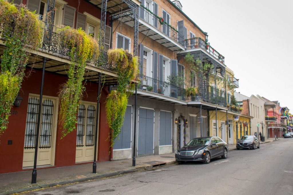 Photo of a street in the French Quarter of New Orleans-a neighborhood which has definitely inspired plenty of New Orleans quotes! There are red and blue buildings on the left, and plants hanging from the balcony.